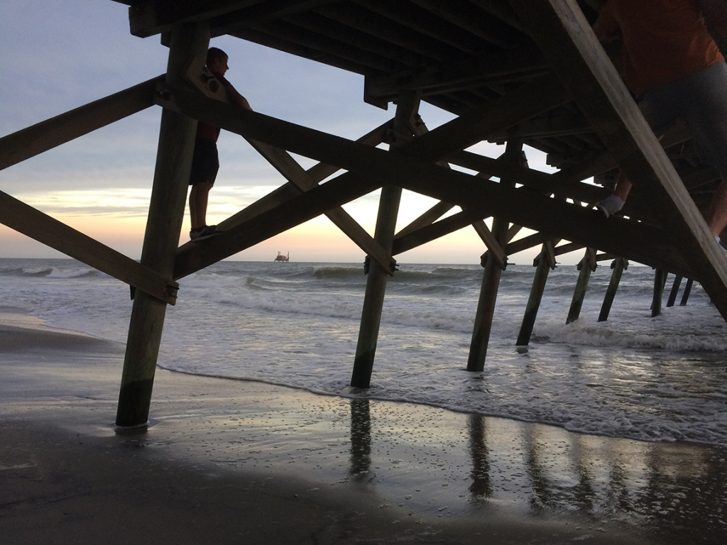 Boys climb timbers of Surfside Beach pier, wrecked in last year's storm. In distance, dredging equipment poised to pump new sand onto beach.